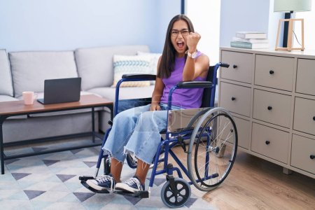 Photo for Young hispanic woman sitting on wheelchair at home angry and mad raising fist frustrated and furious while shouting with anger. rage and aggressive concept. - Royalty Free Image