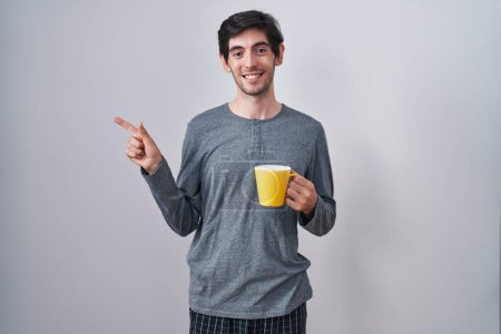 Téléchargez les photos : Jeune homme hispanique portant un pyjama boire une tasse de café gai avec un sourire sur le visage pointant avec la main et le doigt sur le côté avec une expression heureuse et naturelle - en image libre de droit