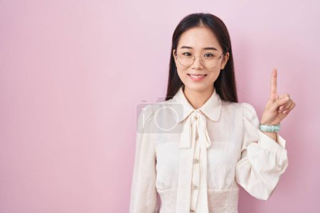 Téléchargez les photos : Jeune femme chinoise debout sur fond rose montrant et pointant vers le haut avec le doigt numéro un tout en souriant confiant et heureux. - en image libre de droit