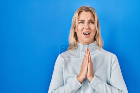 Foto de Mujer caucásica joven de pie sobre fondo azul mendigando y orando con las manos junto con la esperanza expresión en la cara muy emocional y preocupado. mendicidad. - Imagen libre de derechos