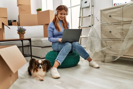 Téléchargez les photos : Jeune femme caucasienne utilisant ordinateur portable et écouteurs assis avec chien à la nouvelle maison - en image libre de droit