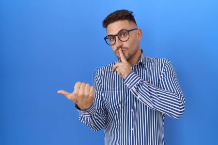 Téléchargez les photos : Homme hispanique à la barbe portant des lunettes demandant à être silencieux avec le doigt sur les lèvres pointant avec la main sur le côté. silence et concept secret. - en image libre de droit