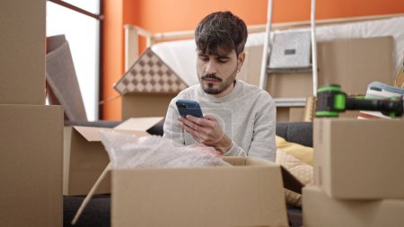 Photo for Young hispanic man unpacking cardboard box using smartphone at new home - Royalty Free Image