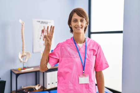 Téléchargez les photos : Femme brune travaillant à la clinique de réadaptation montrant et pointant du doigt le numéro trois tout en souriant confiant et heureux. - en image libre de droit