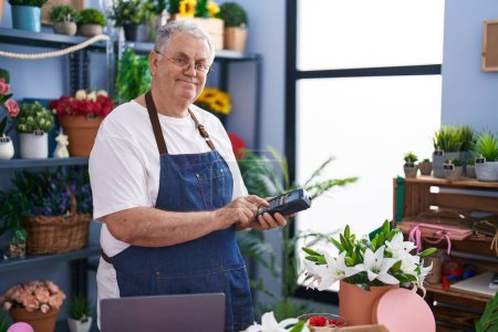 Foto de Hombre de pelo gris de mediana edad florista sonriendo confiado sosteniendo los datos del teléfono en la floristería - Imagen libre de derechos