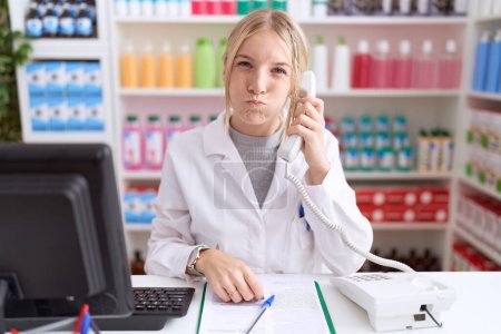 Téléchargez les photos : Jeune femme caucasienne travaillant à la pharmacie pharmacie parlant au téléphone gonflant les joues avec un drôle de visage. bouche gonflée d'air, expression folle. - en image libre de droit