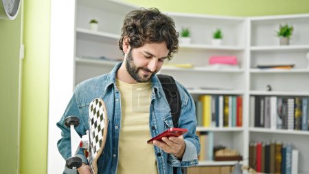 Téléchargez les photos : Jeune homme hispanique étudiant utilisant un smartphone tenant le patin à l'université de la bibliothèque - en image libre de droit