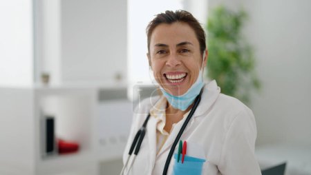 Téléchargez les photos : Femme hispanique d'âge moyen portant un uniforme de médecin et un masque médical à la clinique - en image libre de droit