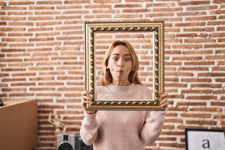 Photo for Hispanic woman holding empty frame making fish face with mouth and squinting eyes, crazy and comical. - Royalty Free Image