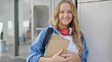 Téléchargez les photos : Jeune étudiante blonde souriante confiante tenant des livres à l'université - en image libre de droit