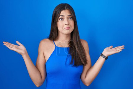 Foto de Hispanic woman standing over blue background clueless and confused expression with arms and hands raised. doubt concept. - Imagen libre de derechos