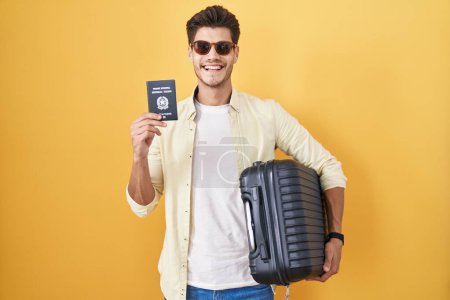 Téléchargez les photos : Jeune homme hispanique tenant une valise allant en vacances d'été tenant un passeport italien souriant avec un sourire heureux et cool sur le visage. montrant les dents. - en image libre de droit