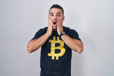 Foto de Joven hombre hispano vistiendo camiseta bitcoin asustado y sorprendido, sorpresa y expresión asombrada con las manos en la cara - Imagen libre de derechos