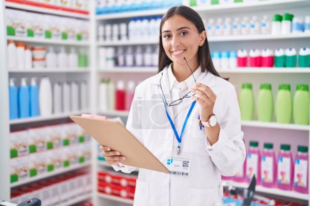 Photo for Young beautiful hispanic woman pharmacist smiling confident holding clipboard and glasses at pharmacy - Royalty Free Image