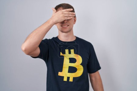Photo for Caucasian blond man wearing bitcoin t shirt smiling and laughing with hand on face covering eyes for surprise. blind concept. - Royalty Free Image