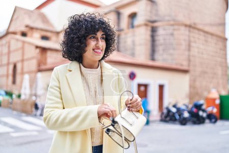 Téléchargez les photos : Jeune femme du Moyen-Orient excutive souriant confiant tenant sac à main à la rue - en image libre de droit