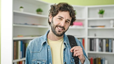 Téléchargez les photos : Jeune homme hispanique étudiant souriant portant avec confiance sac à dos à l'université de la bibliothèque - en image libre de droit
