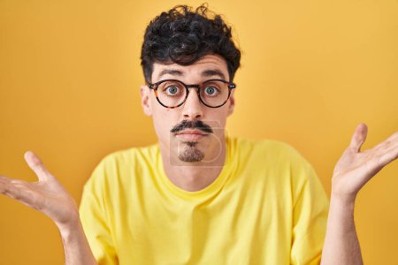 Photo for Hispanic man wearing glasses standing over yellow background clueless and confused expression with arms and hands raised. doubt concept. - Royalty Free Image