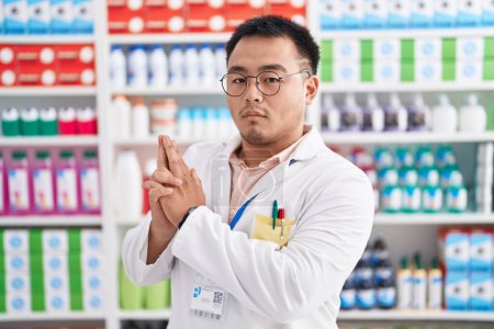 Téléchargez les photos : Jeune homme chinois travaillant à la pharmacie droguerie tenant arme symbolique avec geste de la main, jouer tuer des armes à feu, visage en colère - en image libre de droit