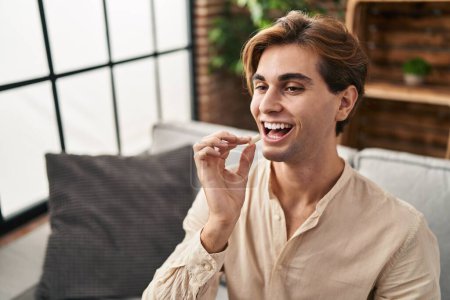 Téléchargez les photos : Jeune homme caucasien tenant chewing-gum assis sur le canapé à la maison - en image libre de droit