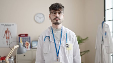 Foto de Joven médico hispano de pie con expresión seria en la clínica - Imagen libre de derechos