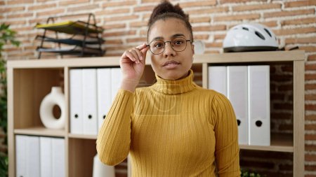 Foto de Joven mujer afroamericana trabajadora de negocios de pie sosteniendo gafas en la oficina - Imagen libre de derechos