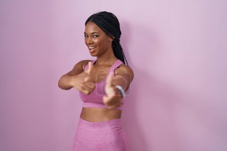 Foto de Mujer afroamericana con trenzas que usan ropa deportiva sobre fondo rosa señalando los dedos a la cámara con la cara feliz y divertida. buena energía y vibraciones. - Imagen libre de derechos