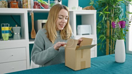 Photo for Young blonde woman unpacking cardboard box at dinning room - Royalty Free Image
