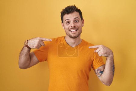 Foto de Joven hombre hispano de pie sobre fondo amarillo mirando confiado con sonrisa en la cara, señalándose con los dedos orgullosos y felices. - Imagen libre de derechos