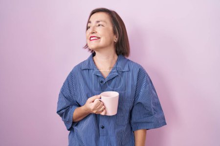 Téléchargez les photos : Femme hispanique du Moyen Âge buvant une tasse de café regardant de côté avec le sourire sur le visage, expression naturelle. riant confiant. - en image libre de droit