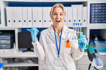 Téléchargez les photos : Jeune femme caucasienne travaillant au laboratoire scientifique tenant des éprouvettes pointant le pouce vers le côté souriant heureux avec la bouche ouverte - en image libre de droit