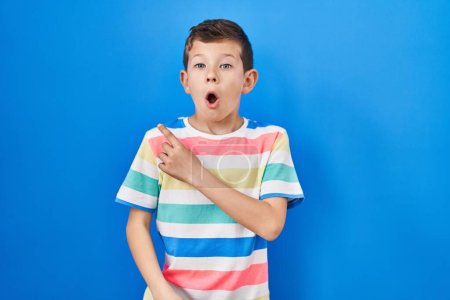 Téléchargez les photos : Jeune enfant caucasien debout sur fond bleu surpris pointant du doigt sur le côté, bouche ouverte expression étonnée. - en image libre de droit