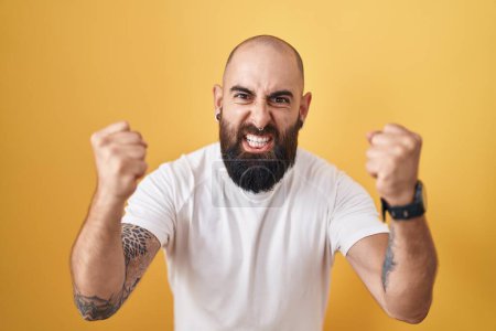 Photo for Young hispanic man with beard and tattoos standing over yellow background angry and mad raising fists frustrated and furious while shouting with anger. rage and aggressive concept. - Royalty Free Image