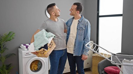Foto de Dos hombres pareja sonriendo confiado celebración cesta con ropa en la lavandería - Imagen libre de derechos