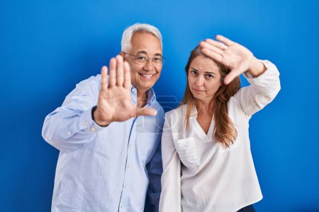 Photo for Middle age hispanic couple standing over blue background doing frame using hands palms and fingers, camera perspective - Royalty Free Image