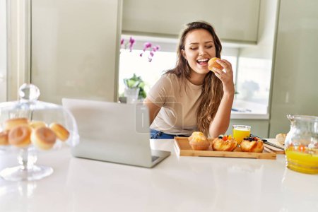 Foto de Joven hermosa mujer hispana desayunando usando laptop en la cocina - Imagen libre de derechos