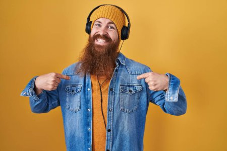 Foto de Hombre caucásico con barba larga escuchando música usando auriculares que se ven confiados con sonrisa en la cara, señalándose con los dedos orgullosos y felices. - Imagen libre de derechos