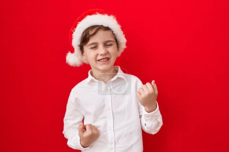 Téléchargez les photos : Petit garçon caucasien portant un chapeau de Noël sur fond rouge célébrant surpris et étonnés pour le succès avec les bras levés et les yeux fermés - en image libre de droit