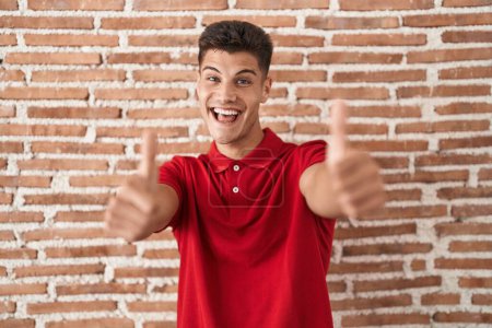 Téléchargez les photos : Jeune homme hispanique debout au-dessus du mur de briques approuvant faire un geste positif avec la main, pouces levés souriant et heureux pour le succès. geste gagnant. - en image libre de droit