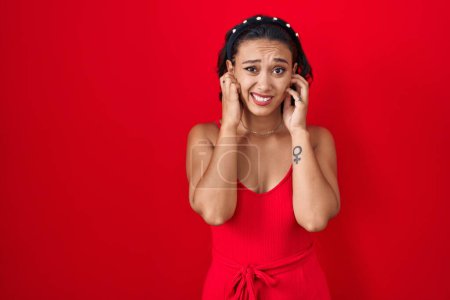 Foto de Mujer hispana joven de pie sobre fondo rojo cubriendo las orejas con los dedos con expresión molesta por el ruido de la música fuerte. concepto de sordo. - Imagen libre de derechos