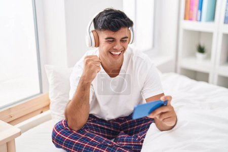 Téléchargez les photos : Jeune homme hispanique souriant confiant jouer au jeu vidéo dans la chambre - en image libre de droit