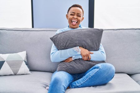 Téléchargez les photos : Femme afro-américaine assise sur le canapé à la maison étreignant oreiller collant la langue heureux avec une expression drôle. - en image libre de droit