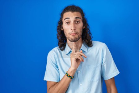 Téléchargez les photos : Jeune homme hispanique debout sur fond bleu pensant concentré sur le doute avec le doigt sur le menton et regardant vers le haut se demandant - en image libre de droit