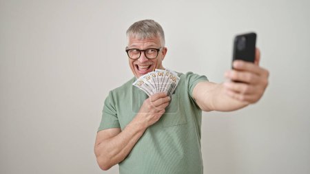 Téléchargez les photos : Moyen Age homme aux cheveux gris prenant selfie par smartphone tenant des dollars sur fond blanc isolé - en image libre de droit