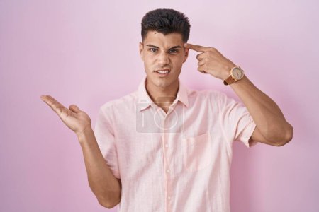 Photo for Young hispanic man standing over pink background confused and annoyed with open palm showing copy space and pointing finger to forehead. think about it. - Royalty Free Image