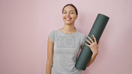 Photo for Young beautiful hispanic woman smiling wearing sportswear holding yoga mat over isolated pink background - Royalty Free Image