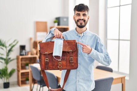 Téléchargez les photos : Jeune homme hispanique avec barbe travaillant au bureau tenant valise et masque de sécurité souriant heureux pointant avec la main et le doigt - en image libre de droit