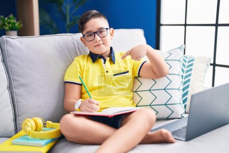 Téléchargez les photos : Jeune enfant hispanique faisant ses devoirs assis sur le canapé à l'air confiant avec le sourire sur le visage, se montrant du doigt fier et heureux. - en image libre de droit