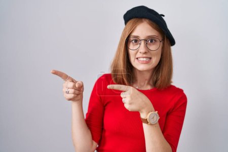 Téléchargez les photos : Jeune rousse femme debout portant des lunettes et béret pointant de côté inquiet et nerveux avec les deux mains, préoccupé et expression surprise - en image libre de droit