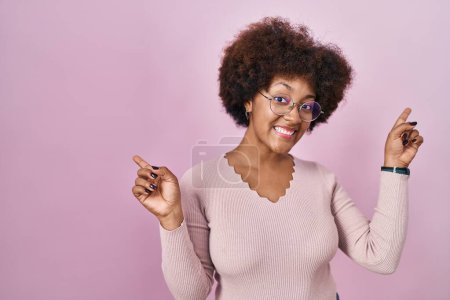 Téléchargez les photos : Jeune femme afro-américaine debout sur fond rose souriant confiant pointant avec les doigts vers différentes directions. espace de copie pour la publicité - en image libre de droit